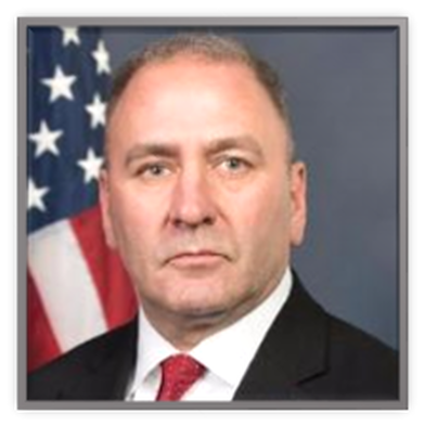 Picture of Representative Clay Higgins in a dark suit, white shirt and red tie with the US flag in the background