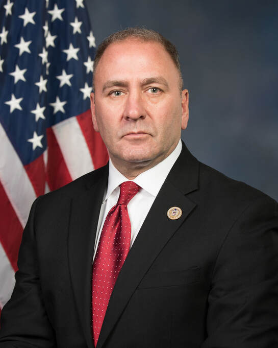 Picture of Representative Clay Higgins in a dark suit, white shirt and red tie with the US flag in the background